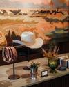 A white cowboy hat, leather belts, a vase with peacock feathers, and small decor items are arranged on a countertop in front of a colorful landscape mural.