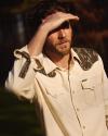 A man wearing a cream shirt with camo-patterned details shields his eyes from the sun with his hand.