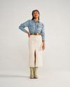 A woman stands against a plain background wearing a light denim shirt, beige ankle-length skirt with front slit, brown belt, and light green boots.