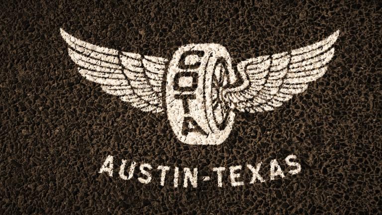 White logo of a tire with wings and "COTA" on it, above the words "AUSTIN-TEXAS," displayed on a textured black surface.