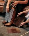 Two people compare leather samples while one wears tall brown cowboy boots with ornate stitching, seated on a leather couch over a patterned rug.