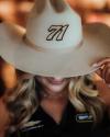 A person with wavy hair wears a wide-brimmed hat embroidered with the number 71, partially covering their face, and a black collared shirt with logos.