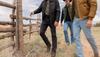 Three men wearing jackets, jeans, and boots walk near a wooden fence in an outdoor rural area with grass and clouds in the sky.