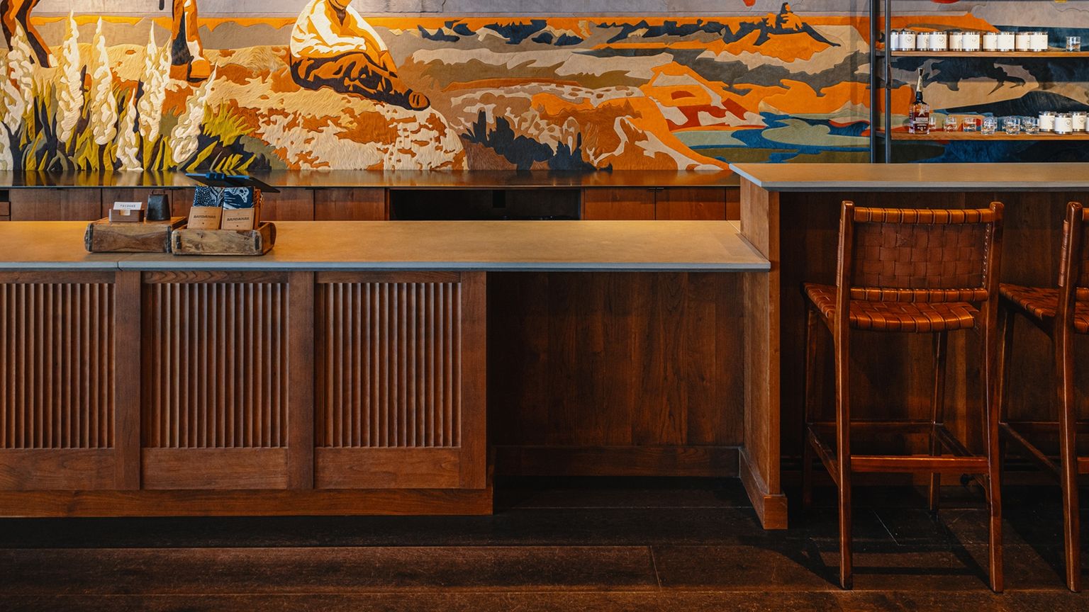 Empty wooden bar with woven chairs, countertop, and a colorful mural on the wall behind, depicting an outdoor scene. Small items are arranged on the shelves in the background.