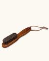 A wooden-handled brush with dense bristles and a leather strap loop at the end of the handle, shown on a plain background.