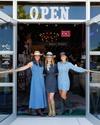 Three women wearing denim and cowboy hats stand in the open doorway of a store with a lit "OPEN" sign above them.