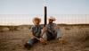 Two people in cowboy hats and denim clothing sit on the ground by a wire fence in a dry, open landscape at sunset.