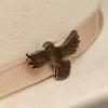 Close-up of a beige felt hat with a matching ribbon band, featuring a bronze eagle-shaped pin attached to the ribbon.