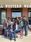 A group of people in cowboy hats pose in front of a store named "Howell Western Wear" with some seated on a bench and others standing behind them.
