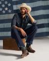 A person in a cowboy hat, denim shirt, and jeans sits on a wooden box in front of a faded American flag backdrop.