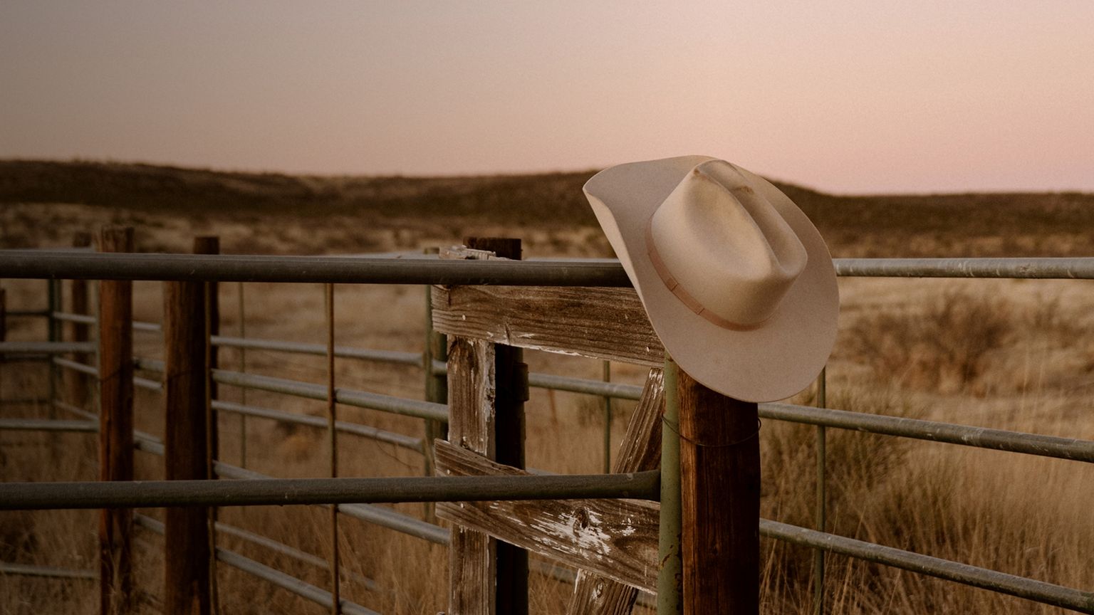 A cowboy hat rests on a wooden fence in a dry, grassy landscape at sunset.