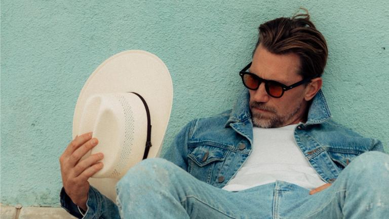 A man in sunglasses and denim clothing sits against a turquoise wall, holding a white hat and looking down at it.