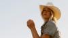 A woman in a light denim jacket and white cowboy hat looks toward the camera against a clear sky.