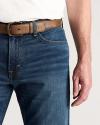 A person wearing a white shirt, blue jeans, and a brown belt is shown from the waist to mid-thigh, with their right arm relaxed at their side.