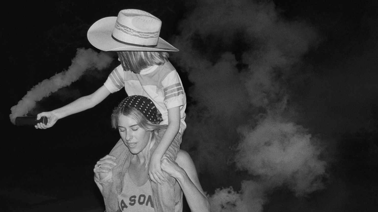 A person carrying a child on their shoulders at night. The child wears a wide-brimmed hat and points an object, with smoke or mist in the background. Black and white photo.