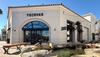 A Tecovas storefront with arched glass doors, a rock-lined pond in front, a wooden bench, and clear sunny skies.