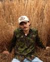 A person wearing a camouflage-patterned jacket and a tan hat sits on dried grass, surrounded by tall, golden-brown grass.