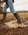 Person wearing jeans and camouflage boots walks through a muddy puddle in a grassy field on an overcast day.