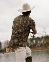 A person wearing a cowboy hat, camouflage shirt, and light pants is fishing by a lake, facing away from the camera. Trees and water are visible in the background.