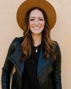 Woman with long brown hair wearing a tan hat, black leather jacket, and black shirt, standing against a light textured wall and smiling at the camera.