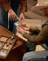 Two people examine ornate silver belt buckles and jewelry displayed in a wooden case. One person wears a cowboy hat and another has tattoos and turquoise jewelry.