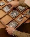 Person organizing a wooden drawer containing ornate silver belt buckles and accessories.