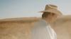 A person wearing a light-colored cowboy hat and shirt stands in a dry, grassy field under a clear sky, facing downward.
