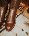 A close-up of brown leather cowboy boots on a patterned rug, with part of a wooden object and a piece of antler visible nearby.