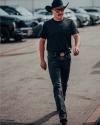 A man wearing a black cowboy hat, sunglasses, a black t-shirt, black jeans, and boots walks confidently in a parking lot.