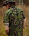 A man wearing a camouflage short-sleeve shirt and matching bucket hat stands outdoors, showing a tattoo on his upper left arm.