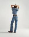 A person stands facing away, wearing a short-sleeved denim shirt, blue jeans, a brown belt, and dark heeled boots, with arms raised and hands resting on their head.