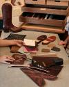 Two people review leather samples, boot pieces, and color swatches on a table, with a finished cowboy boot and an open wooden storage drawer visible in the background.