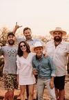A group of eight people outdoors, some wearing hats, smiling and posing together with drinks in hand. Trees and a sunset are in the background.