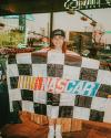A person wearing a NASCAR cap holds a large checkered NASCAR flag inside a store with windows showing street signs and traffic outside.