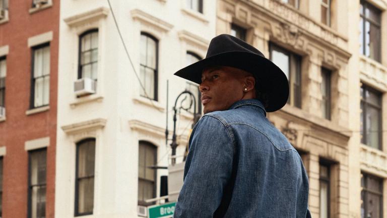 A person in a black cowboy hat and denim jacket stands on a city street with historic buildings in the background.