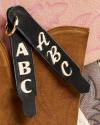 Close-up of two black leather keychains with "ABC" in white letters, resting on brown suede and pink leather surfaces with stitched patterns.
