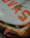 Close-up of the front hood and bumper of a race car with orange lettering, sponsor decals, and a partially visible headlight.