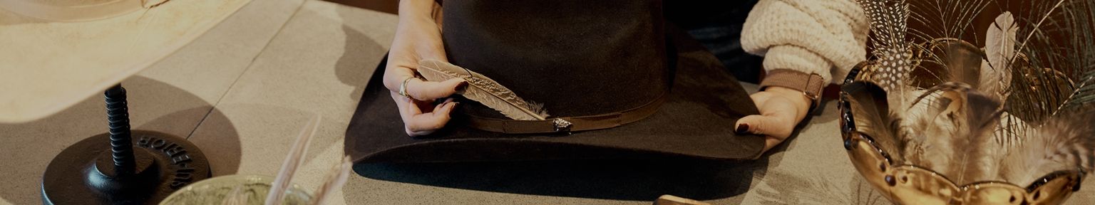 Person decorating a black wide-brimmed hat with a feather, surrounded by various feathers and a lamp on a table.