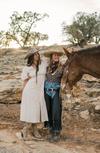 Two people in Western attire stand beside a saddled horse on a rocky terrain with a dog nearby. Trees and a setting sun are in the background.