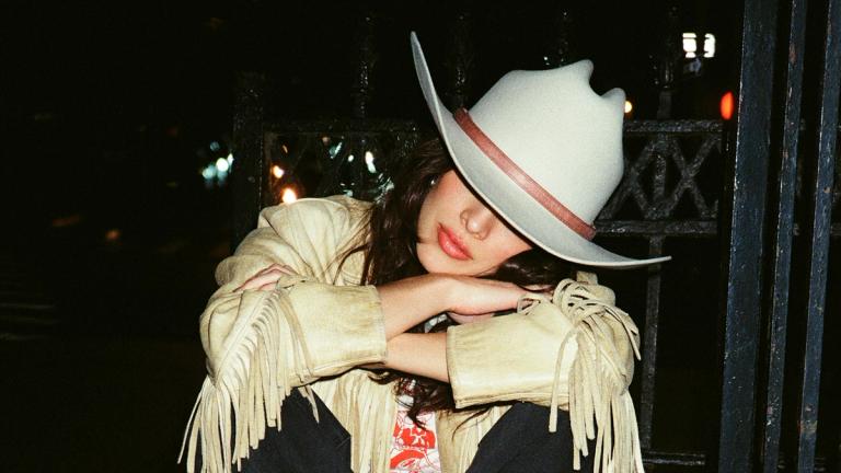 A person wearing a light-colored cowboy hat and fringe jacket sits with arms crossed at night, partially obscuring their face.