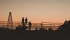 Silhouettes of three people wearing hats sit on a metal fence at sunset, with a windmill and trees in the background.