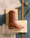 A brown cowboy boot rests on a sketchbook, surrounded by boot design sketches, a pencil, ruler, and a sticky note labeled "Scotch Goat" on a blue grid cutting mat.