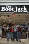 Four people stand in front of The Boot Jack store entrance, smiling, under a sign with "EST. 1976". The store interior is visible behind them.