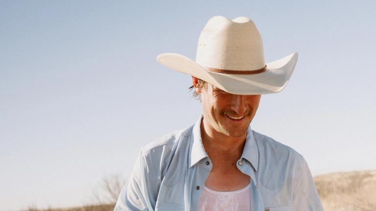 A man wearing a white cowboy hat and light blue shirt stands outdoors under a clear sky, partially smiling with his head tilted down.