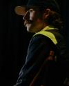 Man wearing a cap and reflective jacket sits in a dark setting, looking to the side with a serious expression.