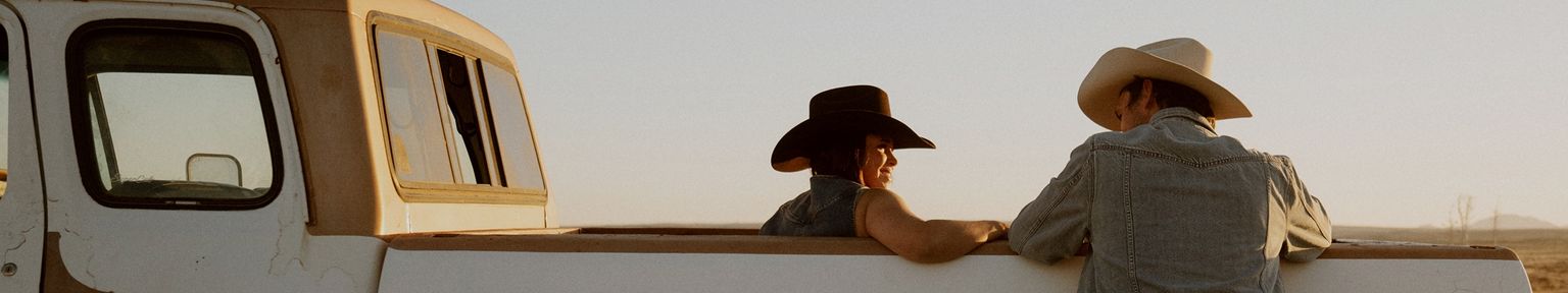 Two people wearing cowboy hats sit in the back of a pickup truck parked in a rural area at sunset, talking and facing each other.