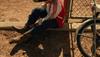 A child in a red and white jacket sits on the ground near a fence, putting on brown boots next to a bicycle.