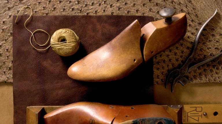 A wooden shoe last, ball of twine, leather sheet, and cobbler’s tools are arranged on a brown work surface.