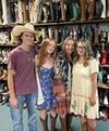 Four people wearing casual outfits and cowboy boots stand in front of shelves filled with various cowboy boots in a store.