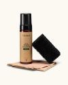 A bottle of Tecovas Hat Foam Cleaner stands next to a black scrubbing pad and a folded beige cloth, all arranged on a light background.